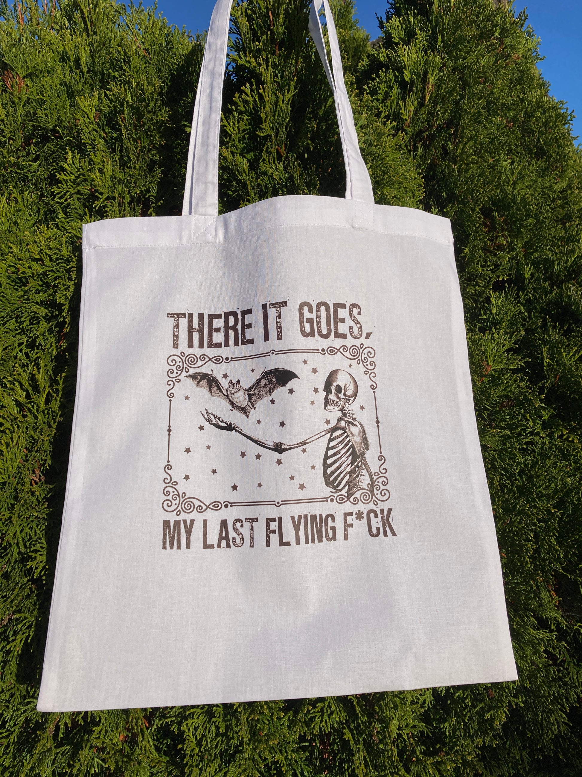 White tote bag with graphic design and text against a green tree background