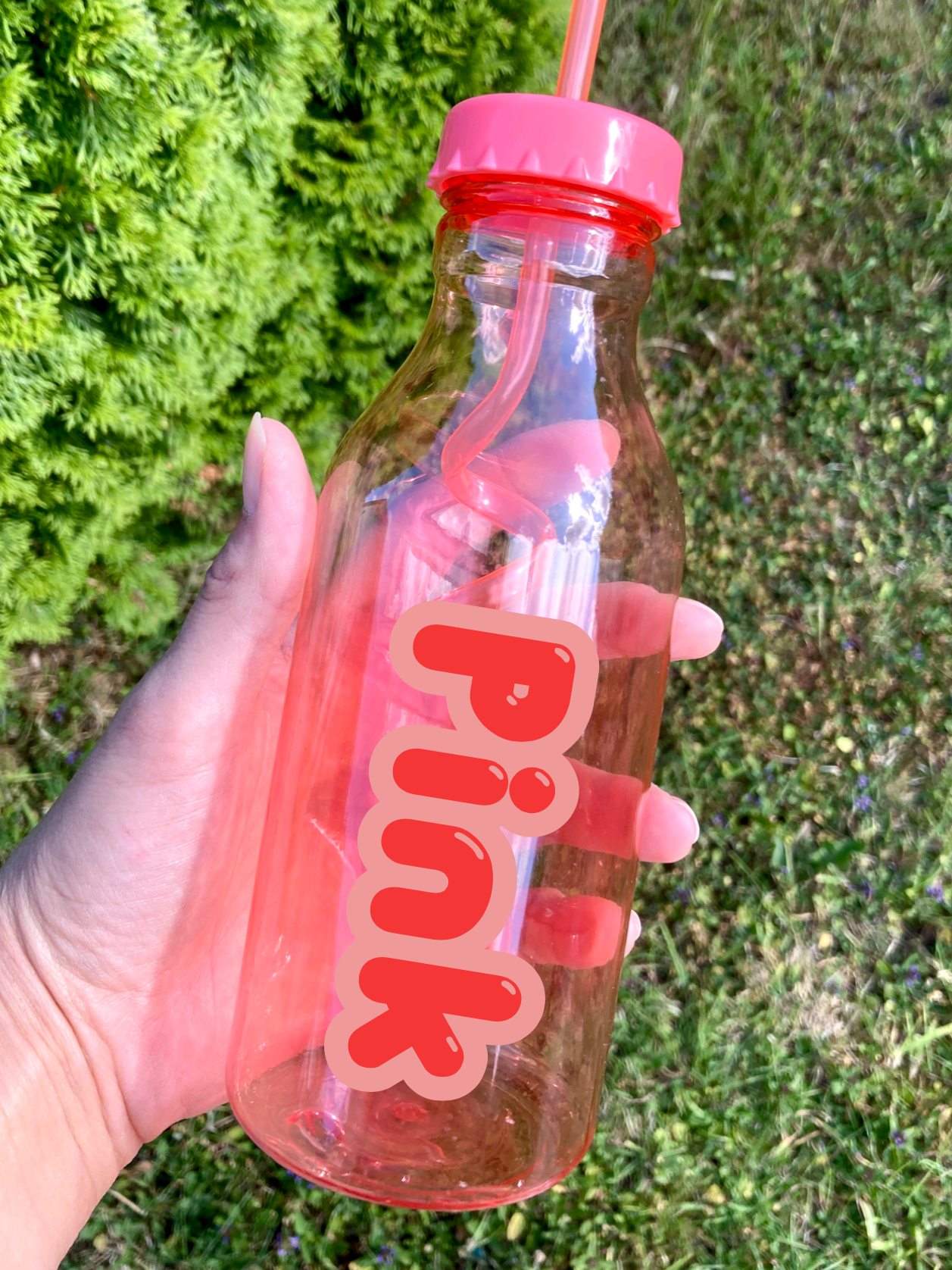 Hand holding a pink water bottle with a straw lid against a grassy background