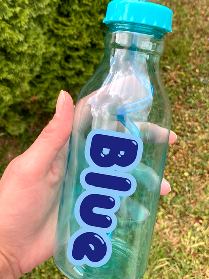 Hand holding a clear water bottle with blue lid and 'Blue' label against a grassy background